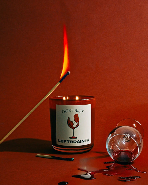 Hand-poured Quiet Riot Candle set against a red background, with a lit match and wine poured nearby, showcasing its rich design and calming home fragrance.