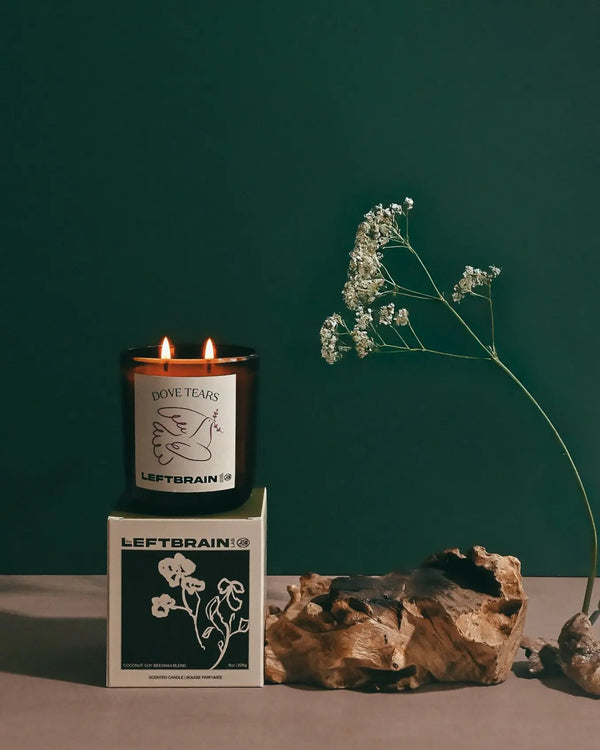 Hand-poured Dove Tears candle posed on its box with a flower and driftwood, showcasing its eucalyptus and fresh flower scent and subtle home fragrance.